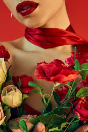 stylisches Frühjahrskonzept, abgeschnittene Ansicht eines jungen Models mit leuchtenden Lippen und Halstuch posiert mit einem Strauß frischer Rosen auf rotem Hintergrund, Modeshooting, Generation z