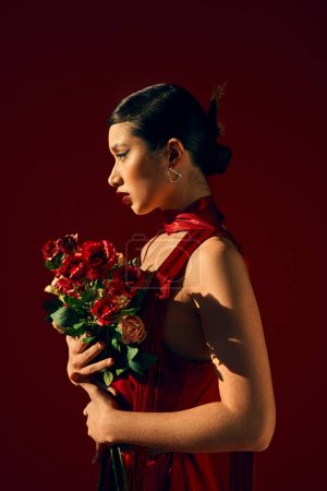 Téléchargez les photos : Vue latérale du modèle asiatique hypnotisant en foulard rouge, avec cheveux bruns et maquillage audacieux posant avec bouquet de roses sur fond sombre, printemps tendance, tournage de mode - en image libre de droit