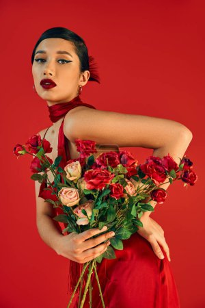 junge und verführerische asiatische Frau mit kühnem Make-up, im trendigen Kleid, das mit der Hand auf Hüfte und Rosen posiert, während sie in die Kamera auf rotem Hintergrund blickt, trendiger Frühling, Modeshooting