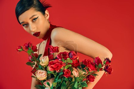 junge und faszinierende asiatische Frau mit kühnem Make-up, brünetten Haaren und verführerischem Blick in die Kamera neben einem Strauß frischer Rosen auf rotem Hintergrund, trendiger Frühling, Generation z
