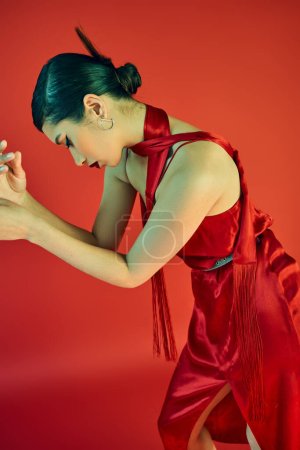 Photo for Stylish asian fashion model with trendy hairstyle and bold makeup posing in elegant dress and neckerchief on red background, trendy spring, charming, glamour, graceful, generation z - Royalty Free Image