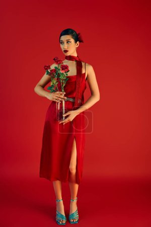 Téléchargez les photos : Pleine longueur de jeune et élégante femme asiatique en robe de sangle élégante et foulard, avec un maquillage audacieux et des cheveux bruns debout avec des roses sur fond rouge, printemps à la mode, génération z - en image libre de droit