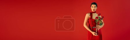 Téléchargez les photos : Jeune et séduisante femme asiatique avec un maquillage audacieux et cheveux bruns, dans une robe élégante et un foulard poising avec bouquet de roses sur fond rouge, photographie de mode de printemps, bannière - en image libre de droit