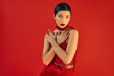 young asian woman with brunette hair and bold makeup holding hands on chest and looking away while posing in neckerchief and dress on red background, fashionable spring