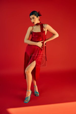full length of attractive asian woman in elegant dress, neckerchief and turquoise sandals holding hands on hip while standing on red background with lighting, gen z fashion, trendy spring concept