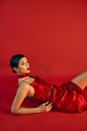 moda primavera, generación z, mujer asiática joven con pelo morena y maquillaje en negrita que pone sobre fondo rojo, pañuelo, vestido de correa elegante, maquillaje en negrita, expresivo, glamour 