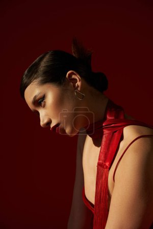 Téléchargez les photos : Profil de jeune femme asiatique avec cheveux bruns, maquillage audacieux et boucle d'oreille argentée posant en foulard et robe de sangle sur fond rouge foncé avec éclairage, concept de mode de printemps - en image libre de droit
