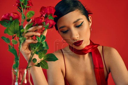 sinnliche und träumerische asiatische Frau mit kühnem Make-up, brünetten Haaren und stylischem Halstuch, das frische Rosen auf rotem Hintergrund berührt, Frühlingsstil, Modefotografie