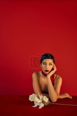Femme asiatique coûteuse en foulard élégant, avec des cheveux bruns et un maquillage audacieux assis à table près de l'orchidée, tenant la main près du visage et regardant loin sur fond rouge, concept de printemps à la mode