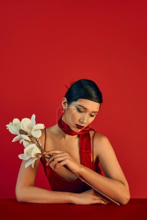 youthful fashion, trendy spring concept, charming asian woman in neckerchief and strap dress sitting at table with white blooming orchid on red background