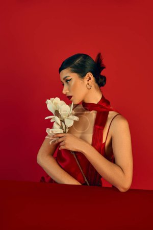 Foto de Concepto de moda de primavera, mujer asiática joven sentada en la mesa con orquídea blanca y mirando hacia otro lado sobre fondo rojo, cabello moreno, maquillaje audaz, vestido de correa, pañuelo, estilo z gen - Imagen libre de derechos