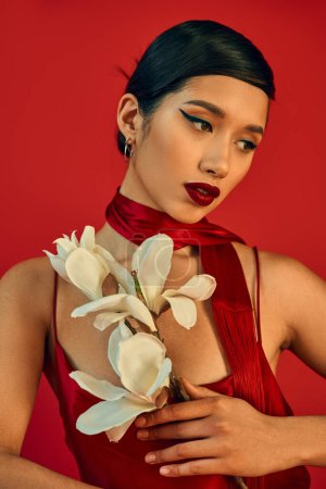 Porträt der schönen asiatischen Frau in elegantem Trägerkleid, Halstuch, mit kühnem Make-up und brünetten Haaren, die weiße blühende Orchidee auf rotem Hintergrund halten, Gen-Z-Mode, stilvolles Frühjahrskonzept