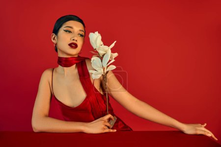 Foto de Mujer asiática elegante y encantadora en vestido elegante y pañuelo, con pelo moreno y maquillaje audaz mirando la orquídea floreciente cerca de la mesa sobre fondo rojo, fotografía de moda de primavera - Imagen libre de derechos