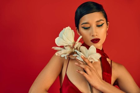 mujer asiática elegante y tierna con cabello moreno, maquillaje audaz, en vestido de correa elegante y pañuelo posando con orquídea blanca en flor sobre fondo rojo, concepto de primavera de moda