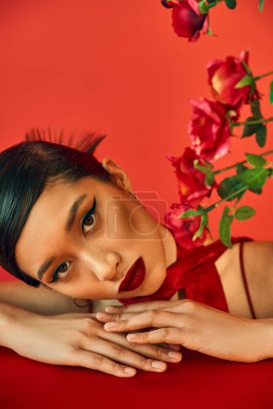 Porträt eines jungen und attraktiven asiatischen Models mit kühnem Make-up und brünetten Haaren, das in die Kamera blickt, während es auf einem Tisch in der Nähe von Rosen auf rotem Hintergrund liegt, stilvolles Frühjahr, Modeshooting
