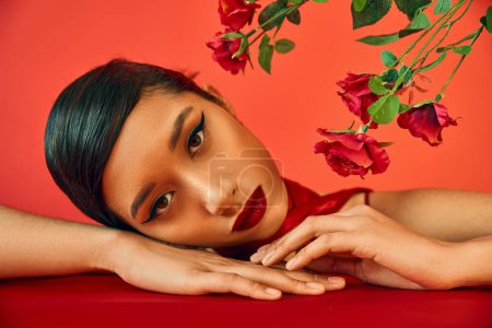 portrait of charming and sensual asian woman with brunette hair and bold makeup laying on table near fresh roses and looking at camera on red background, youthful fashion, trendy spring