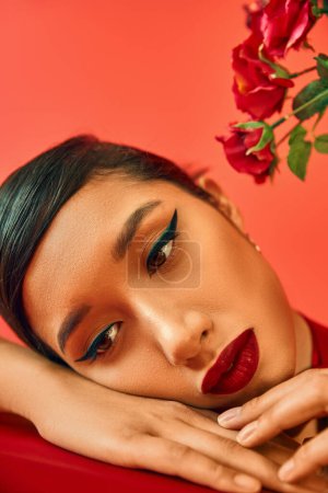 close up portrait of mesmerizing and pensive asian woman with bright makeup and brunette hair dreaming with hands near face next to flowers on red background, spring style, generation z