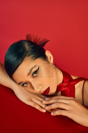 spring fashion concept, portrait of young and beautiful asian fashion model leaning on table and looking at camera on red background, brunette hair, bold makeup, expressive gaze, generation z