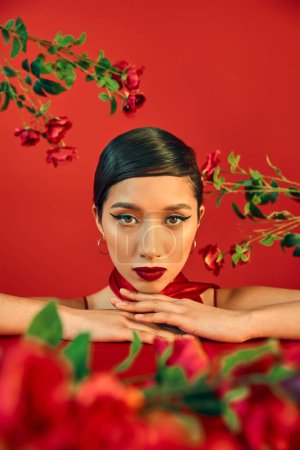 Foto de Retrato de mujer asiática joven y encantadora con maquillaje brillante y cabello moreno mirando a la cámara rodeada de rosas frescas sobre fondo rojo, concepto de fotografía de moda de primavera - Imagen libre de derechos