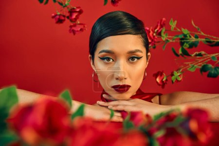 gen z fashion, Porträt einer charmanten und jungen asiatischen Frau mit hellem Make-up, die zwischen frischen Rosen auf Rot in die Kamera blickt, Frühlingsmodefotografie, verschwommener Vordergrund