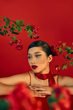 portrait of stylish and beautiful asian woman with bold makeup, brunette hair and neckerchief leaning on table and looking away near roses on red, blurred foreground