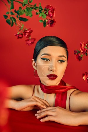 Foto de Retrato de fascinante y soñadora mujer asiática posando cerca de rosas frescas sobre fondo rojo, maquillaje audaz, cabello moreno, elegante pañuelo, concepto de primavera de moda - Imagen libre de derechos