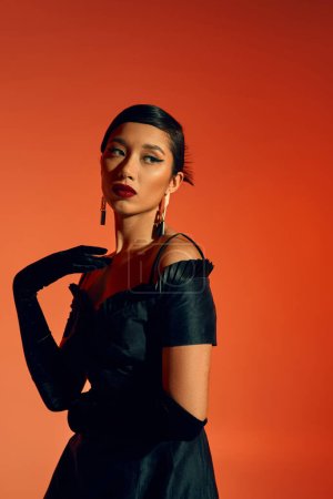 Photo for Stylish and attractive asian fashion model in black cocktail dress, long gloves and trendy earrings touching shoulder and looking away on red and orange background, spring fashion photography - Royalty Free Image