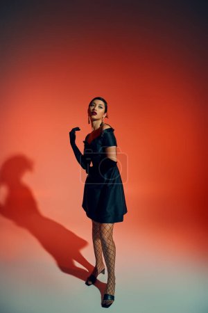 full length of glamour and youthful asian woman in black gloves, fishnet tights and cocktail dress standing in expressive pose on red background with lighting effects, trendy spring concept