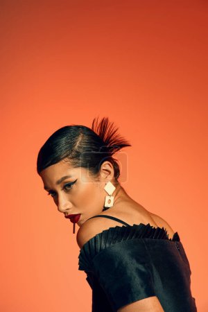 Porträt einer glamourösen asiatischen Frau mit fettem Make-up und brünetten Haaren, die trendige Ohrringe und ein schwarzes elegantes Kleid trägt, während sie auf orangefarbenem Hintergrund posiert, Generation Z, Frühlingsmodefotografie