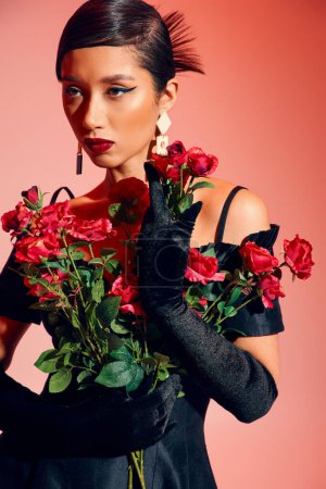 mujer asiática encantadora y de moda con maquillaje audaz, en pendientes de moda, guantes largos y vestido de cóctel negro posando con rosas rojas y frescas sobre fondo rosa, generación z, concepto de primavera de moda