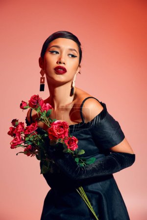 Photo for Young and expressive asian woman with brunette hair, bold makeup, in black cocktail dress, long gloves and trendy earrings holding roses and looking away on pink background, stylish spring concept - Royalty Free Image