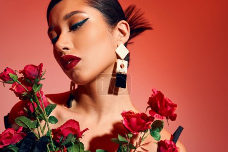 Porträt einer attraktiven und modischen Asiatin mit brünetten Haaren, fettem Make-up, trendiger Frisur und Ohrringen, die mit roten und frischen Rosen auf rosa und rotem Hintergrund posiert, Frühjahrsmodekonzept