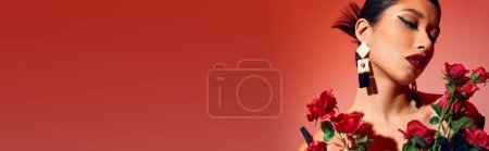 portrait of pretty and sensual asian woman with brunette hair, bold makeup, trendy earrings and hairstyle posing near fresh roses on pink and red background, gen z fashion, stylish spring, banner
