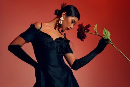trendy spring, expressive and youthful asian woman with brunette hair and trendy earrings, wearing long gloves and black dress, posing with hand on hip and burgundy peony on red and pink background
