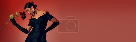 Foto de Mujer asiática joven y elegante en pendientes de moda, guantes largos negros y elegante vestido de cóctel posando con la mano en la cadera y peonía de color burdeos sobre fondo rojo y rosa, concepto de estilo primavera, bandera - Imagen libre de derechos
