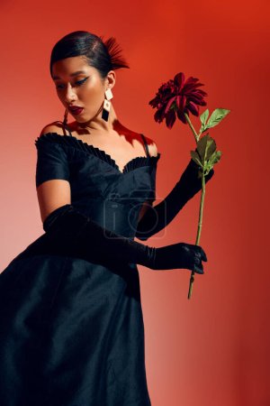 Foto de Elegante concepto de moda de primavera, mujer asiática joven en vestido de cóctel negro, guantes largos y pendientes de moda posando con peonía de color burdeos sobre fondo rojo y rosa, generación z - Imagen libre de derechos