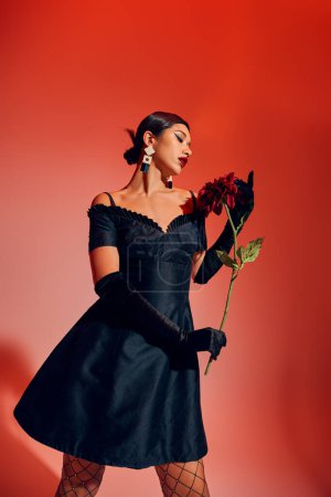 mesmerizing asian woman with brunette hair and bold makeup, wearing black cocktail dress and long gloves holding burgundy peony on red and pink background, trendy spring concept