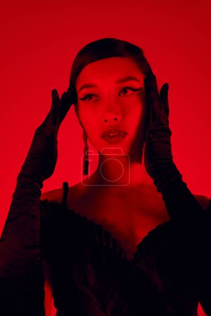 Foto de Retrato de impresionante mujer asiática con cabello moreno y maquillaje audaz cogidas de la mano cerca de la cara y mirando hacia otro lado sobre un fondo vibrante con efecto de iluminación roja, vestido negro, guantes largos, primavera de moda - Imagen libre de derechos