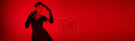 mujer asiática joven y de moda en vestido de cóctel negro y guantes largos posando sobre un fondo vibrante con efecto de iluminación roja y espacio de copia, fotografía de moda de primavera, pancarta