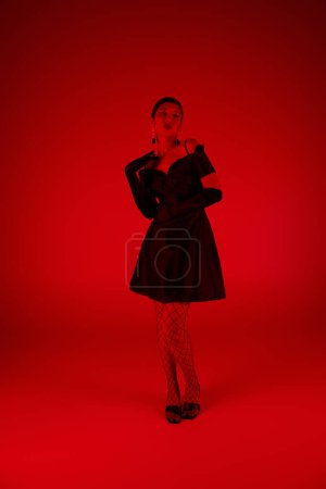 fotografía de moda de primavera, longitud completa de la mujer asiática con estilo en vestido de cóctel negro, medias de rejilla y guantes largos de pie sobre un fondo vibrante con efecto de iluminación roja, generación z