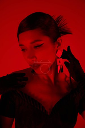 Photo for Portrait of charming asian woman with brunette hair and bold makeup, in black dress and long gloves touching trendy earring on vibrant background with red lighting effect, spring fashion concept - Royalty Free Image