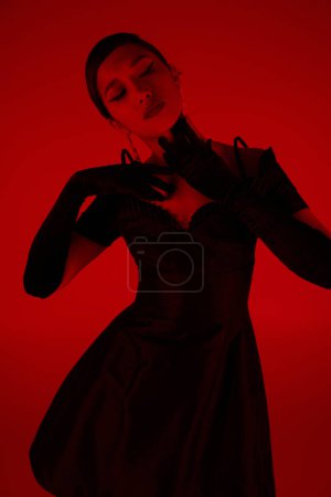 expressive and graceful asian woman in black long gloves and elegant cocktail dress touching neck and posing on vibrant background with red lighting effect, spring fashion photography