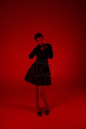Foto de Longitud completa de joven y elegante modelo asiático posando en vestido de cóctel negro, guantes largos y medias de rejilla sobre un fondo vibrante con efecto de iluminación roja, primavera de moda, sesión de fotos de moda - Imagen libre de derechos