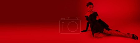 Foto de Longitud completa de la mujer asiática de moda en vestido de cóctel, guantes largos negros y medias de rejilla sentado y mirando hacia otro lado en un fondo vibrante con efecto de iluminación roja, concepto de moda de primavera, bandera - Imagen libre de derechos