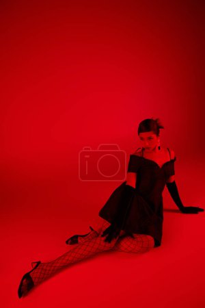 generation z, spring fashion concept, full length of young asian woman in black dress, long gloves and fishnet tights sitting on vibrant background with red lighting effect