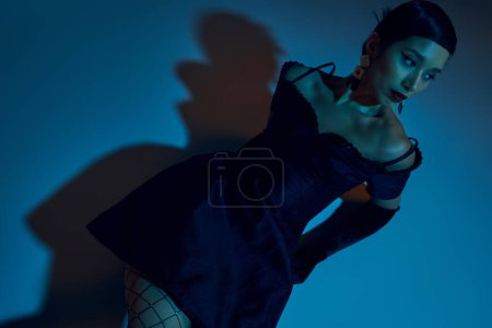 Foto de Vista de ángulo alto de la atractiva mujer asiática con cabello moreno, en vestido de cóctel negro y guantes largos mirando hacia otro lado mientras está sentado sobre fondo azul con efecto de iluminación cian - Imagen libre de derechos