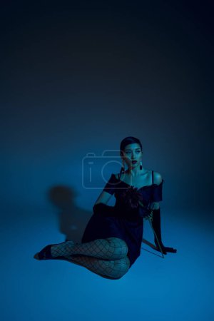 in voller Länge ansprechende asiatische Frau in schwarzem Cocktailkleid, Netzstrumpfhose und langen Handschuhen, die wegschauen, während sie auf blauem Hintergrund mit Cyan-Lichteffekt sitzt, Modemodel 