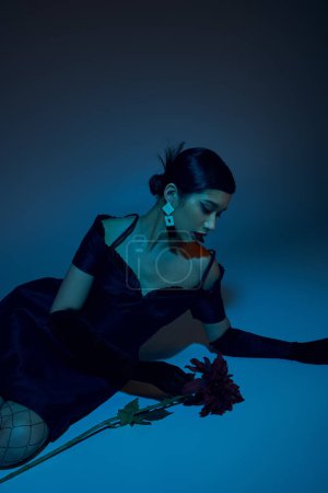 appealing asian woman with brunette hair and trendy earrings, in black cocktail dress and long gloves sitting near peony flower on blue background with cyan lighting effect, spring fashion concept