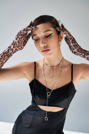 elegant asian woman with brunette hair and bold makeup, wearing animal print gloves, black strap dress and silver accessories, holding hands near head on grey background, spring fashion photography