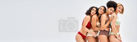 Smiling and multiethnic women in colorful lingerie looking at camera while hugging each other isolated on grey, different body types and self-acceptance concept, multicultural models, banner 
